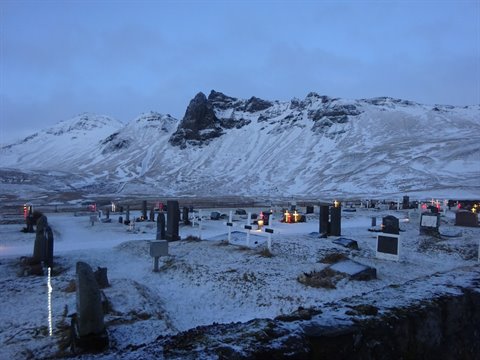 Snowy graveyard with headstones and lights before rugged mountains, twilight light
