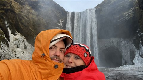 Couple in orange and red jackets take a selfie in front of an icy waterfall