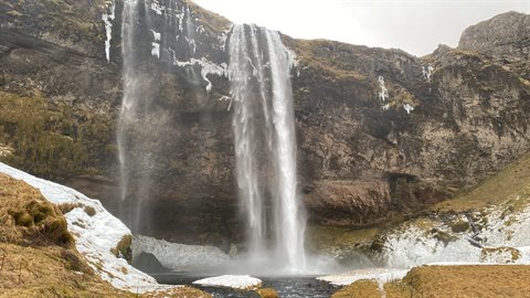 Waterfall falling over dark cliffs with snow, diffuse light, water splashing into the pool