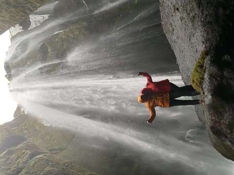 Traveler in yellow and red jackets beneath a waterfall, mossy rock, soft light