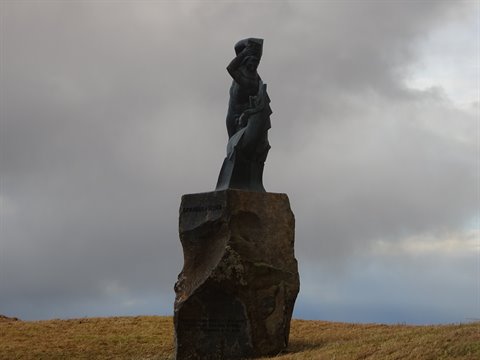 Dark stone statue on boulder, yellow fields under cloudy sky