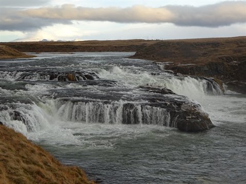 Wide waterfall over volcanic rocks, yellow grass, cloudy sky, soft light, flowing water