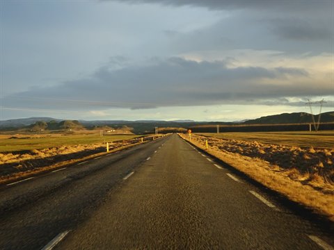 Straight asphalt road through yellow fields and hills, cloudy evening light