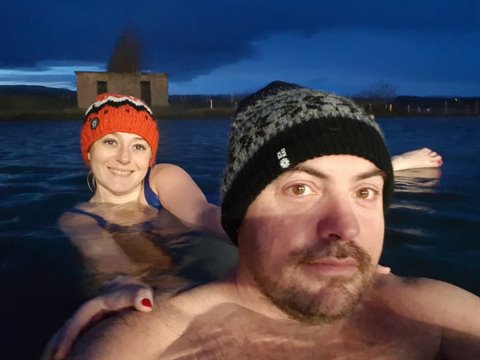 Couple in wool hats soaking in warm water at night, building behind, blue light