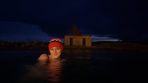 Woman in orange hat swimming in night hot springs, small stone hut, dark cloudy sky