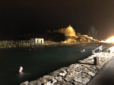 Swimmers in a nighttime hot spring pool, stone wall, warm light on hill and trees