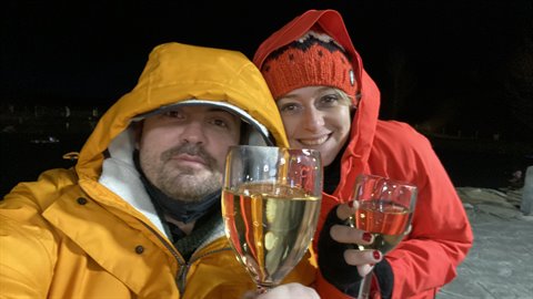 Couple in colorful jackets holding wine glasses, nighttime outdoor selfie