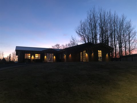 Wooden lodge lit at dusk, lawn in front, bare trees behind