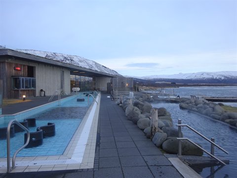 Outdoor thermal pool with wooden panels, lit lamps, snowy mountains, cold light