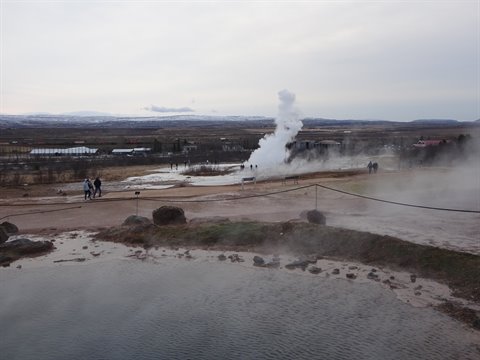 Geysers erupting white steam, visitors on sandy path, diffused light