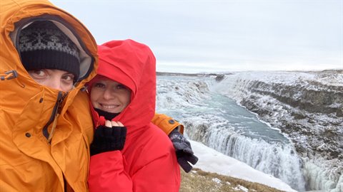 Two travelers in orange and red jackets selfie by an icy waterfall, soft daylight