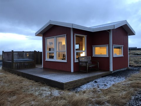 Red wooden cabin lit at dawn, wooden deck, bench and frosty grass fields