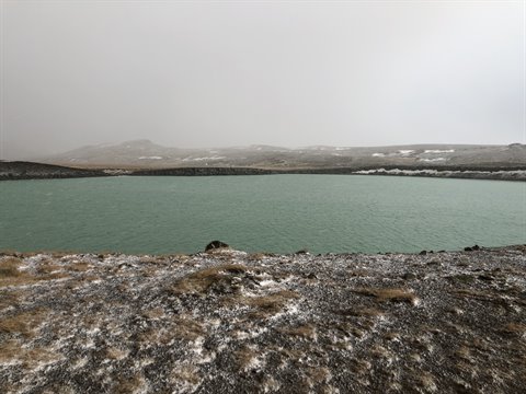 Green lake surrounded by rocky snowy ground under grey sky