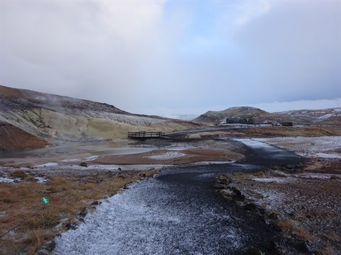 Snow-dusted path toward wooden boardwalk among steaming volcanic hills, cold diffuse light