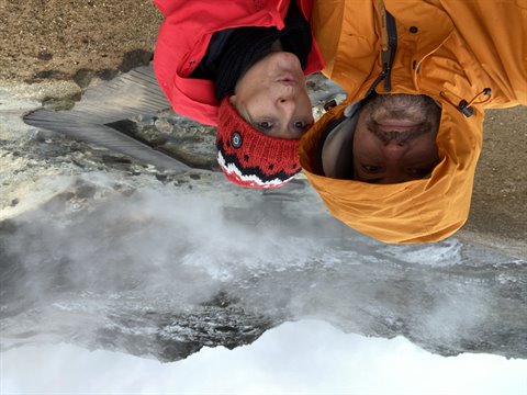 Two hikers in orange and red jackets above steaming geothermal ground