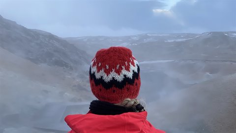 Traveler in red coat and knitted wool hat watches steam rising over misty hills