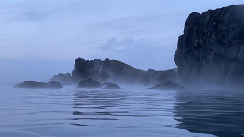 Dark cliffs shrouded in mist above calm blue sea, soft diffuse light