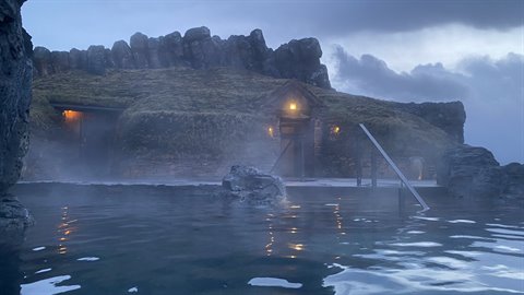 Outdoor thermal pool in front of a turf-roofed house among rocks, warm lights at dusk