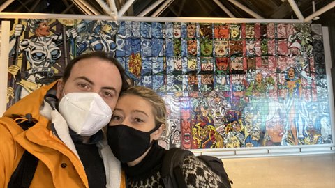 Masked couple takes a selfie in front of a comic mosaic in a station
