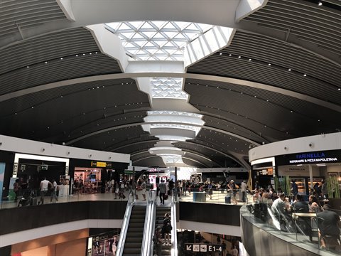 Travelers among shops and escalators under an airport's curved ceiling, natural light from skylights