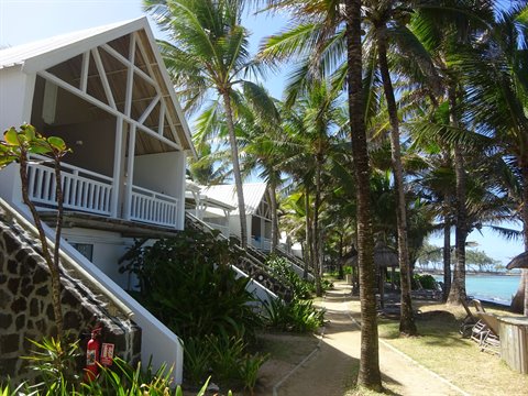 White wooden bungalow with balcony among palm trees, sandy path toward the sea, sunlight