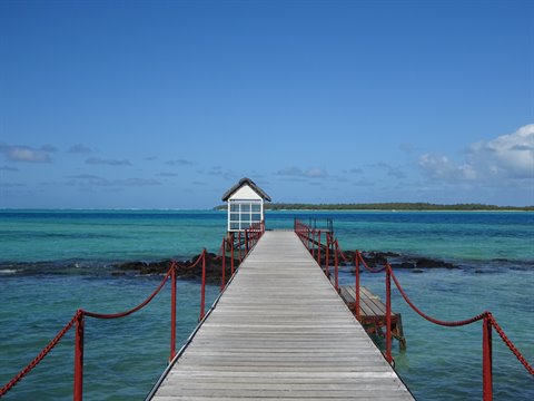 Wooden jetty with red ropes leading to white hut over turquoise lagoon, blue sky