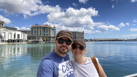 Couple in caps and sunglasses taking a selfie in front of waterfront buildings, cloudy sky