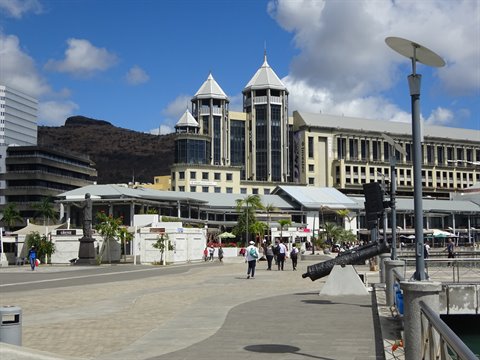 Waterfront square with historic cannon, pedestrians and cone-topped towers under clouds