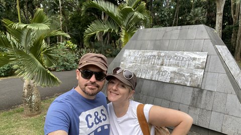 Couple in caps and sunglasses taking a selfie in front of a grey stone monument surrounded by tropical palms