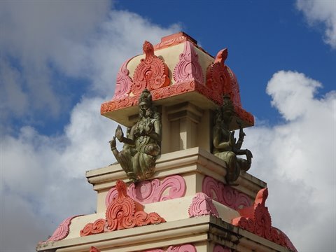 Deity statues on a pink and orange painted temple spire against cloudy sky