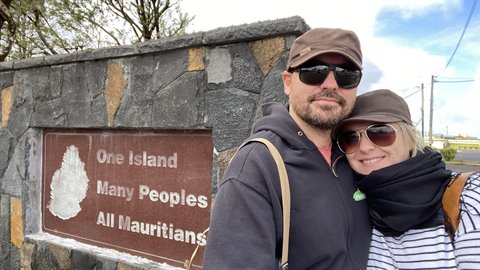 Couple wearing caps and sunglasses beside a stone sign with engraved map, cloudy day