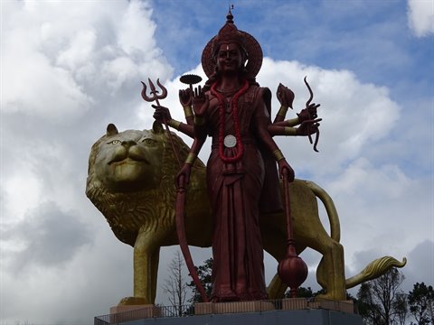 Red metal statue of multi-armed deity on golden lion, cloudy sky