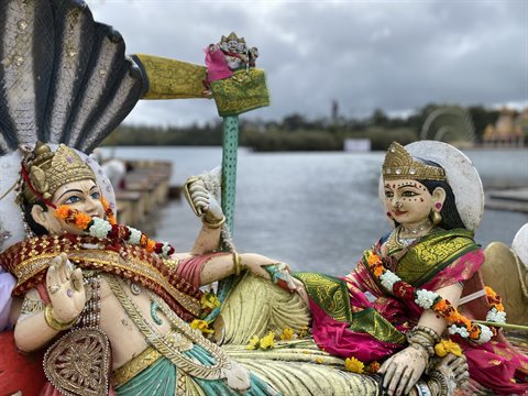 Decorated Hindu deity statues with garlands beside a lake under cloudy sky