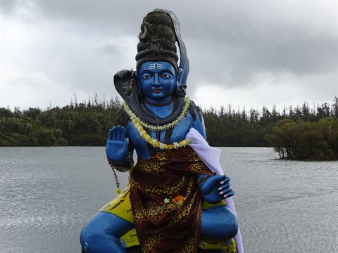 Blue Hindu deity statue with flower garland beside a lake under cloudy skies