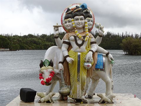 Marble statue of multi-faced deity beside sacred cows, lake and trees behind, cloudy light