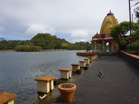 Yellow Hindu temple by the lakeside, paved promenade, green hills, cloudy sky