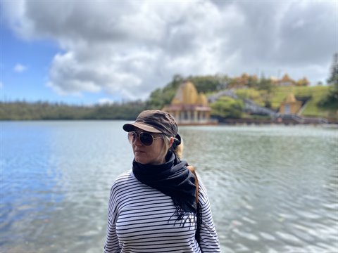 Woman wearing cap, sunglasses and scarf by a lake, temple on hill, cloudy light