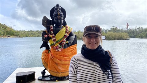 Smiling woman beside statue of deity with garlands, lake and trees, soft light