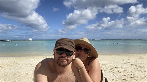 Couple in hat and sunglasses hugging on sand, turquoise sea with boats