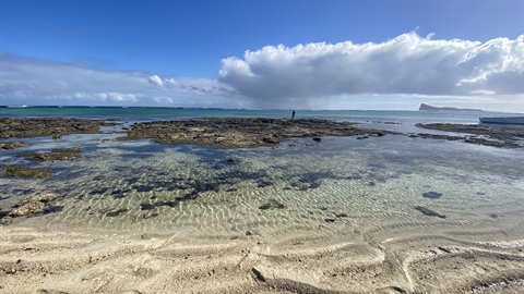 Person walking on exposed rocks in front of low sandy tide, cloudy sky
