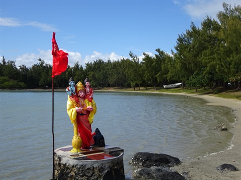 Colorful religious statue on stone pedestal by tree-lined bay, bright sunlight