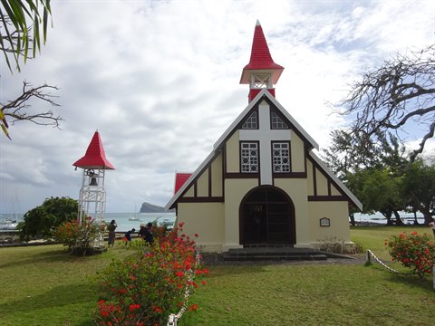 Small church with red roof facing the sea, cloudy daylight, flowers and bell tower