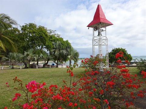 White bell tower with red roof among red flowers and palms, cloudy light