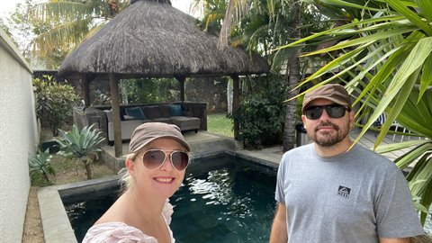 Smiling couple wearing hats beside tropical pool, thatched gazebo in background, daylight