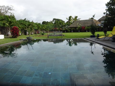 Empty tiled swimming pool reflects the cloudy sky, tropical garden and thatched roof