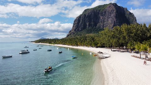 Boats in turquoise water approaching white sand beach beneath green rocky mountain