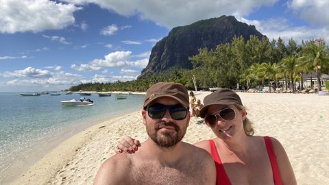 Couple takes a selfie on the beach, mountain and boats behind, bright sunlight