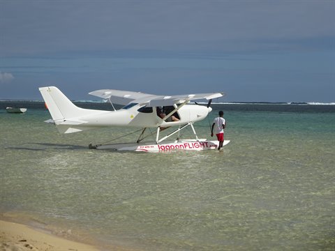 White seaplane resting in shallow turquoise water, a man walking beside it, natural light
