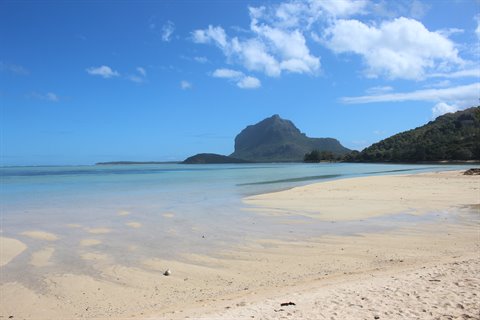 Pale sandy beach and turquoise sea under white clouds, green mountain in background