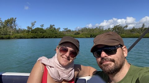 Couple in caps and sunglasses takes a selfie on a boat over turquoise water, tree-lined shore behind, bright sun